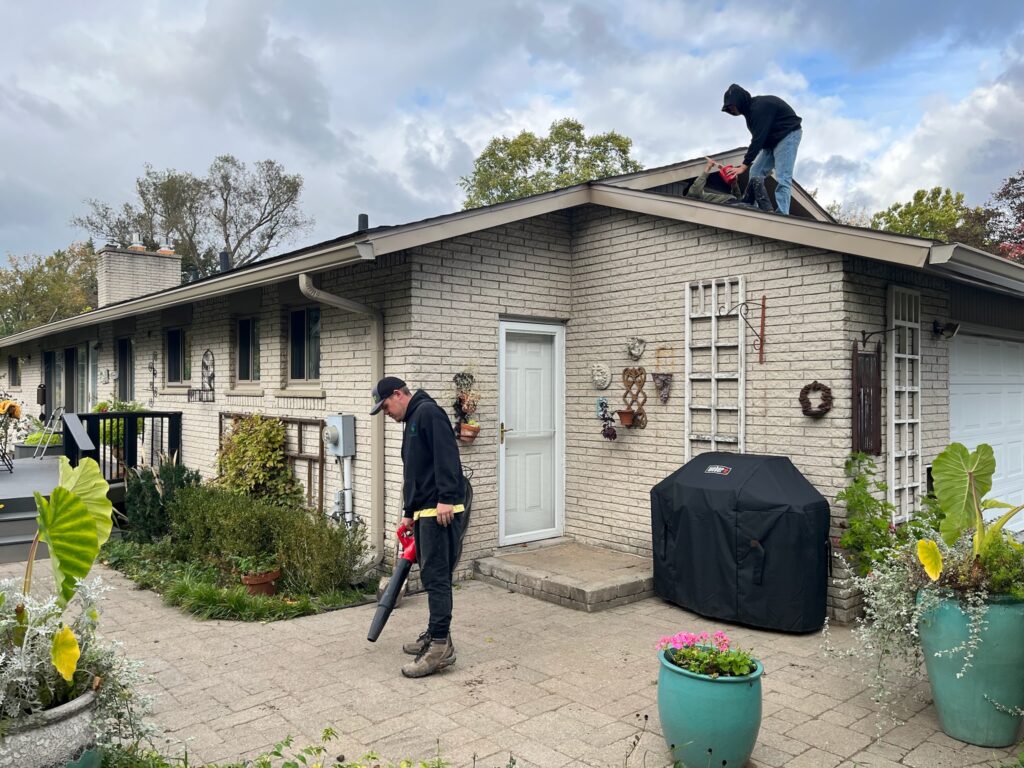 two men working on a beige brick home roof and a man holding a leaf blower